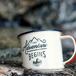 Tazza da campeggio in metallo smaltato bianco con bordo blu e stampa decorativa "The Adventure Begins", appoggiata su una roccia.