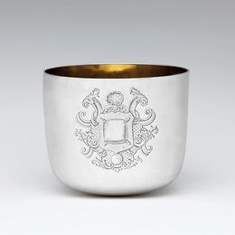 Bicchiere senza manico in metallo argentato, con interno dorato e stemma decorativo inciso al centro del fronte, su fondo grigio chiaro.